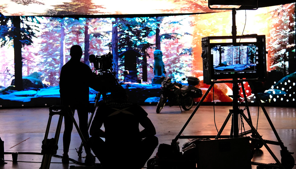 Virtual movie set. POV is standing behind the director and camera person, with the digital monitor to the right, which is projected onto a panorama screen behind the actors