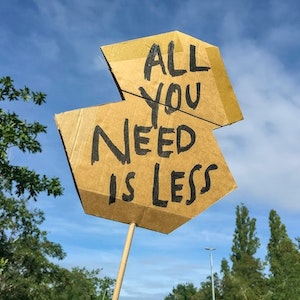 Cardboard sign that says "All you need is less"
