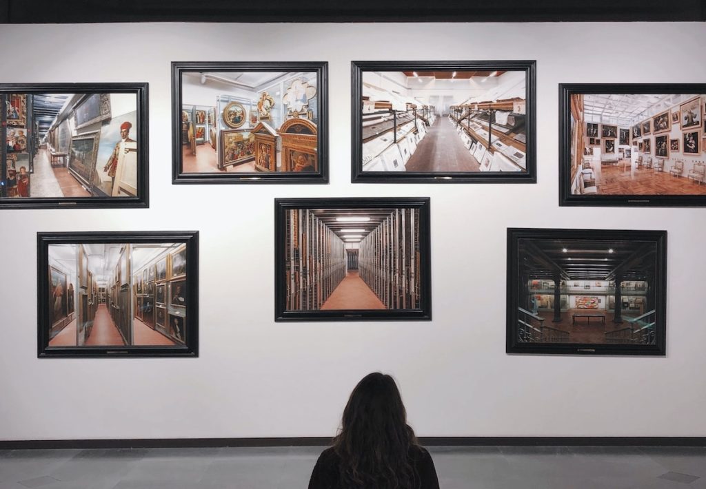 Woman in black-and-white sweater sitting on the museum floor looking up at seven photographs