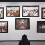 Woman in black-and-white sweater sitting on the museum floor looking up at seven photographs