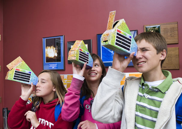 Three middle school kids holding up house-shaped boxes to their faces and looking through a hole toward the ceiling.