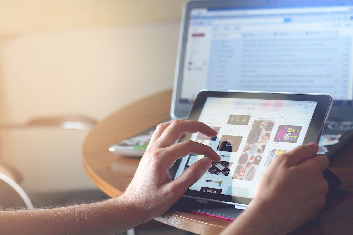 A person is using their hands to scroll through images on a tablet while a laptop sits in the background