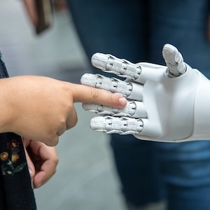 A girl reaches out her finger to touch the small, white plastic hand of a robot.