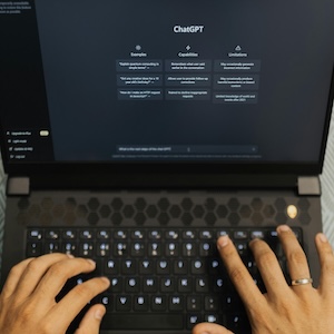 View looking down at a laptop's keyboard, with a person's hands typing into ChatGPT.